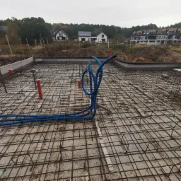 Zbrojenie płyty fundamentowej z wystającymi rurami instalacyjnymi w kolorze pomarańczowym i niebieskim, widok na budynki w tle.