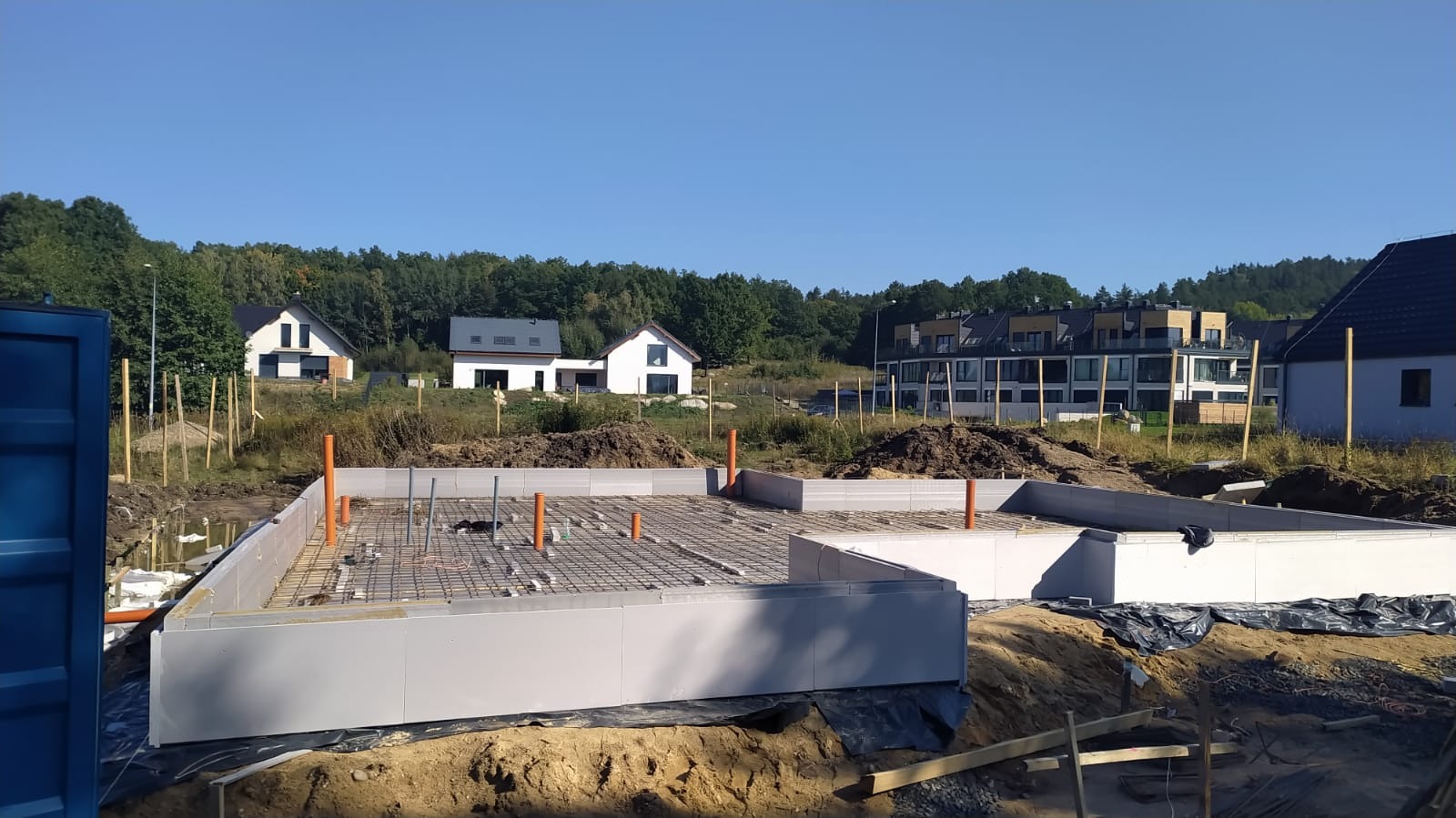 Wylany fundament z widoczną siatką zbrojeniową, rurami instalacyjnymi i szalunkiem w otoczeniu placu budowy z widokiem na domy jednorodzinne w tle.