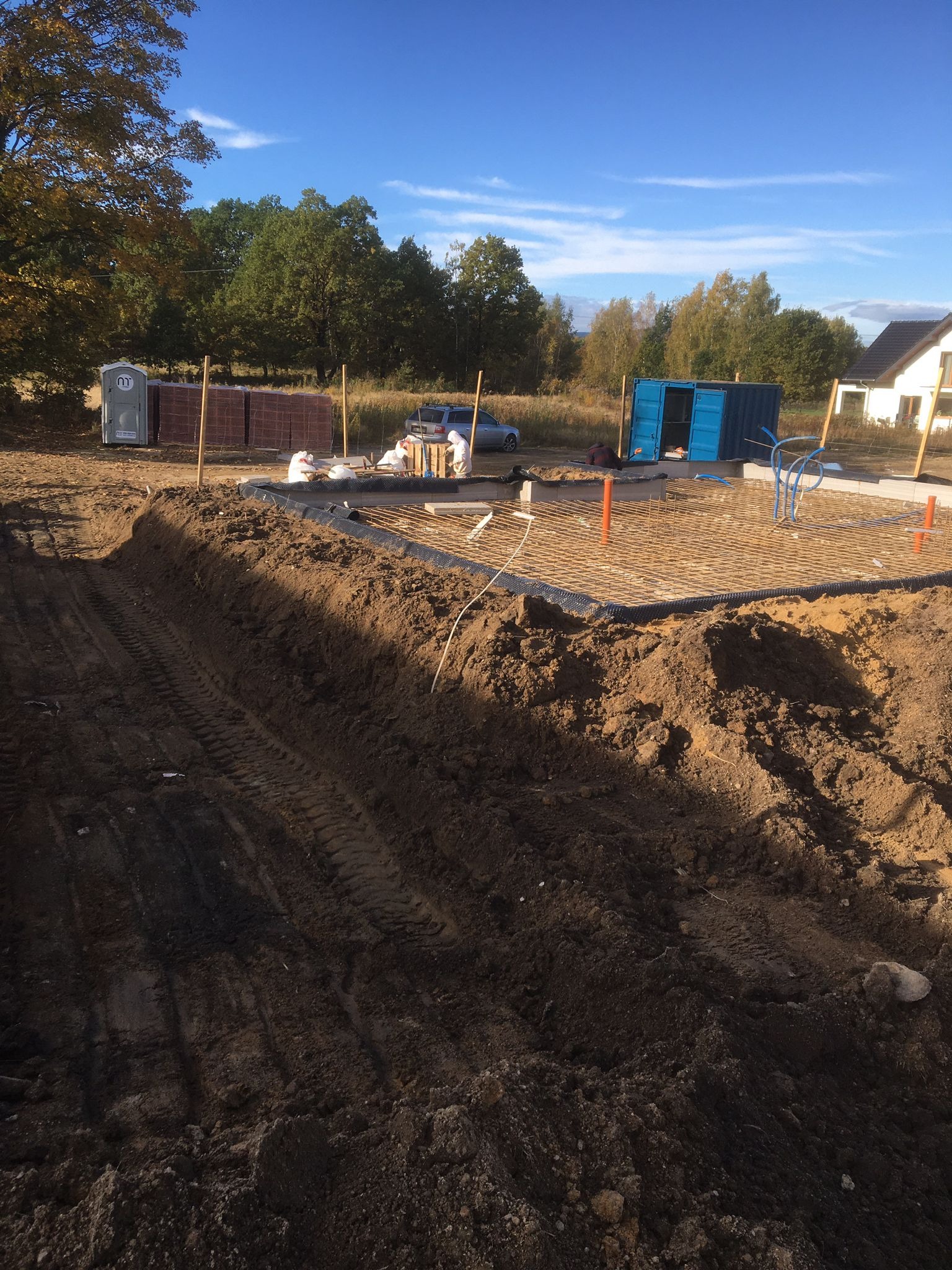 Przygotowanie terenu pod budowę: wykop z widocznymi śladami opon, stalowa siatka zbrojeniowa na fundamencie, palety cegieł i przenośna toaleta w tle.