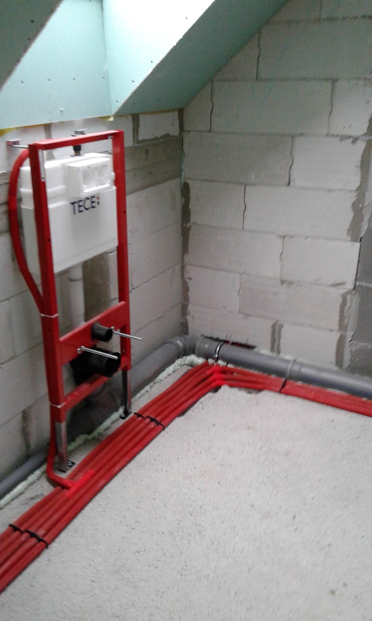 Montaż stelaża podtynkowego TECE z czerwonymi rurami instalacji wodnej w surowym wnętrzu z bloków betonowych i skośnym sufitem z płyty gipsowej.