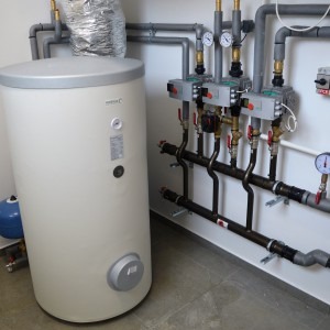 Biały, pionowy zbiornik na wodę w kotłowni, z widocznymi rurami, zaworami i manometrami, zamontowany na kafelkach w jasnym pomieszczeniu.