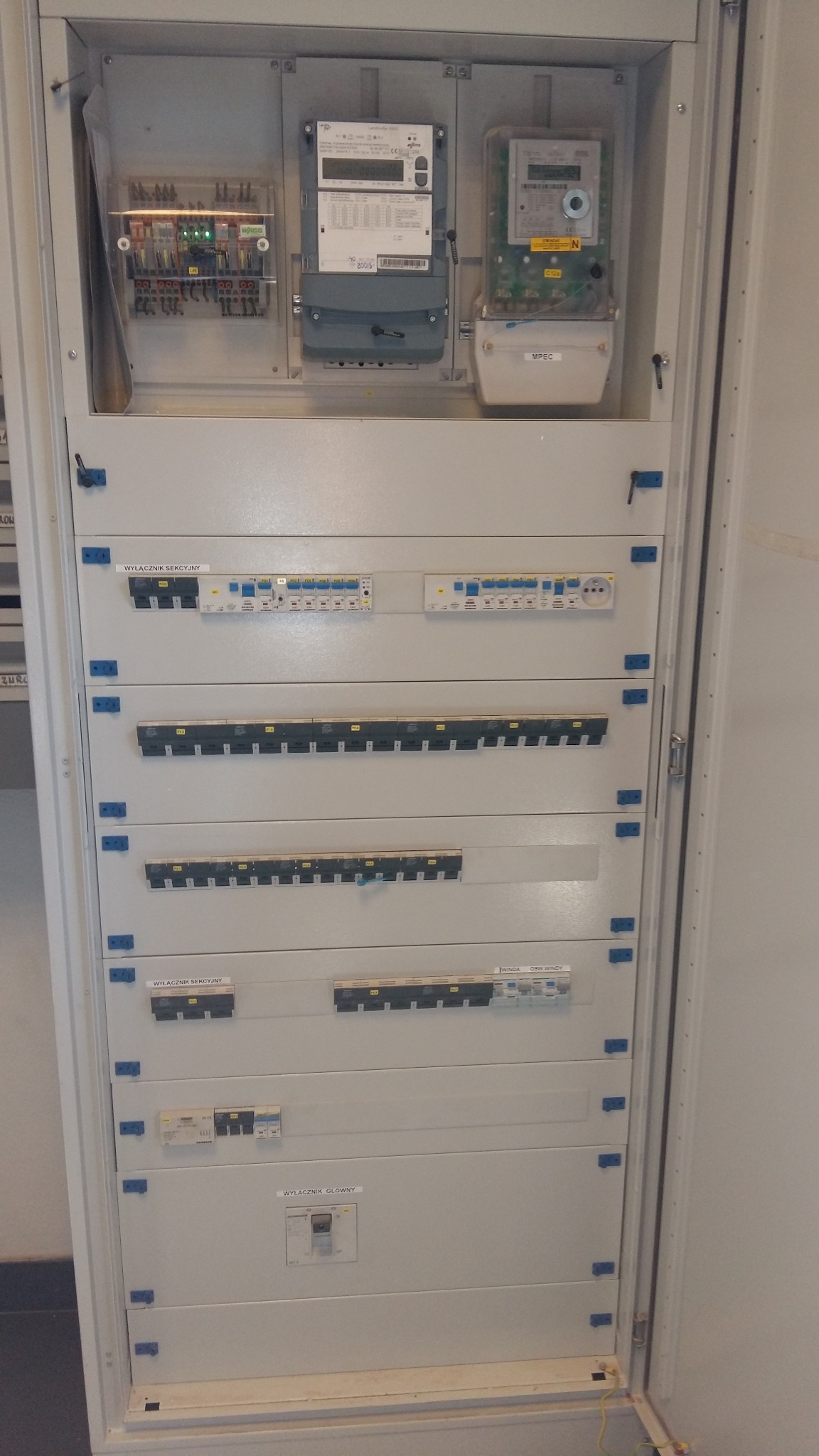 Otwarta szafa elektryczna z licznikami i zabezpieczeniami różnicowoprądowymi, widoczne bezpieczniki sekcyjne i wyłącznik główny, elementy opisane etykietami.