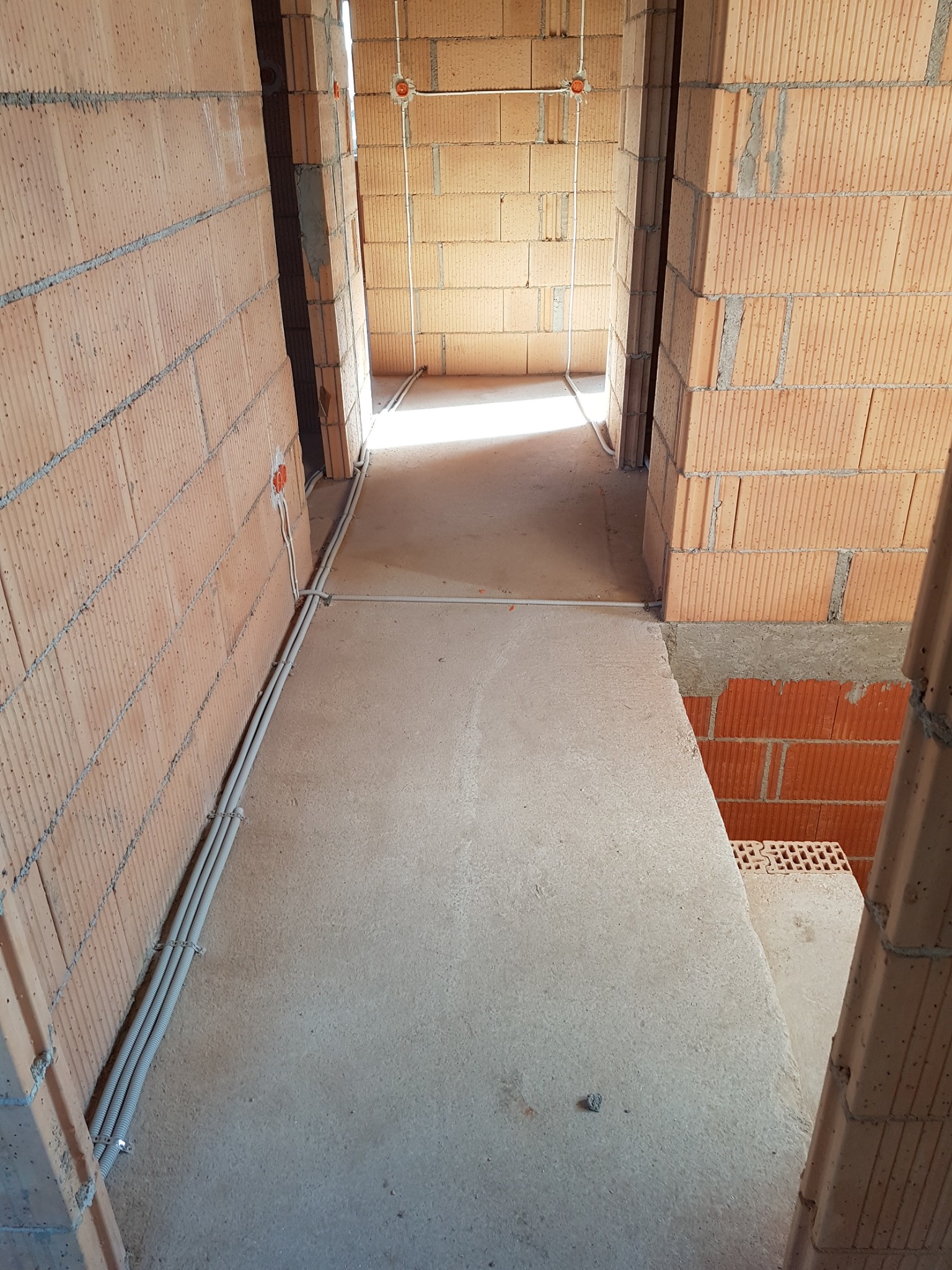 Wnętrze w stanie surowym w Opolu: widoczne ściany z cegły, betonowa podłoga i rury instalacji elektrycznej prowadzone wzdłuż ściany.