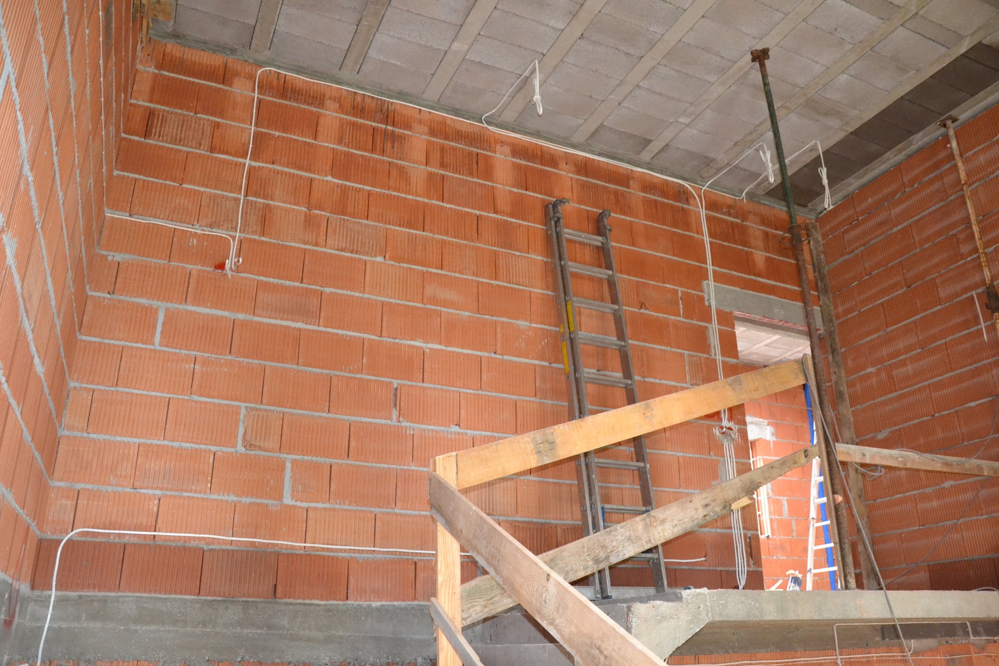 Wnętrze domu w trakcie budowy: ściany z czerwonej cegły, częściowo położone kable elektryczne, drabina oparta o ścianę, drewniane szalunki schodów i podpory stropu.