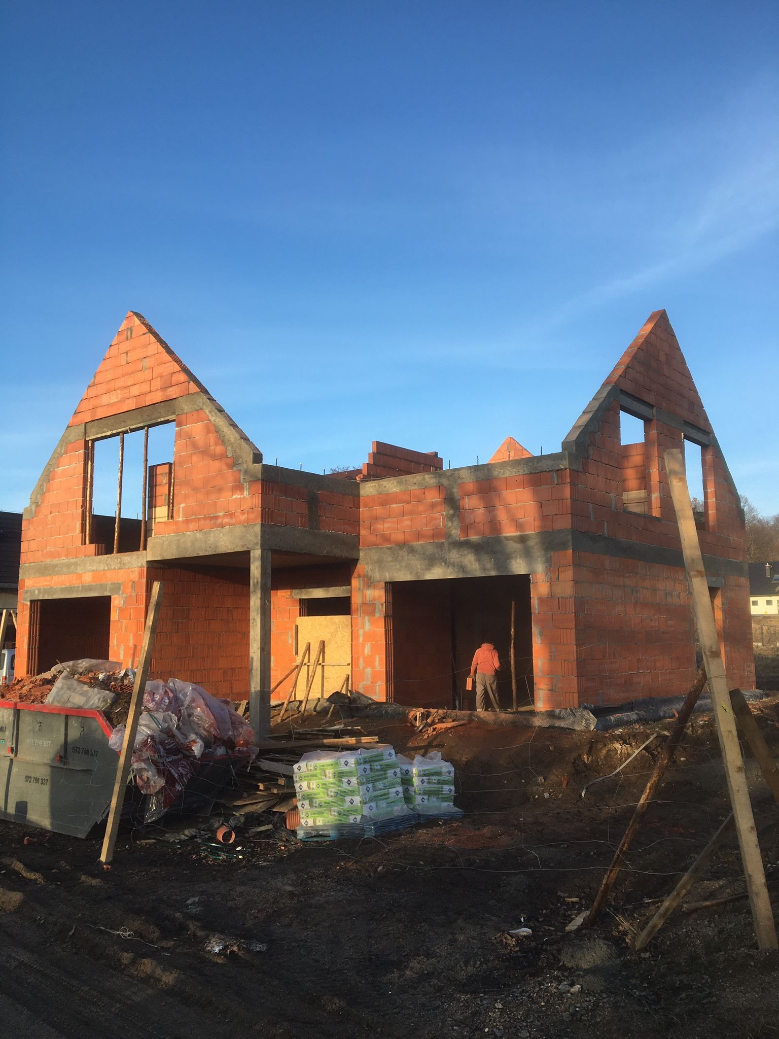 Wznoszenie dwukondygnacyjnego budynku z czerwonej cegły, widoczny szkielet konstrukcji, okna bez ram, pracownik wewnątrz, palety z materiałami budowlanymi i kontener na gruz przed budynkiem.