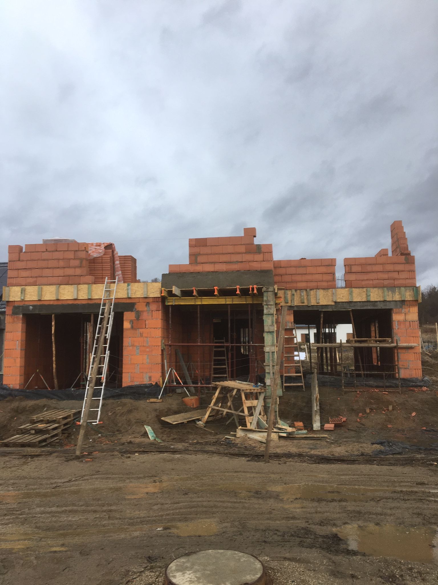Wznoszenie ścian parterowego domu z czerwonej cegły ceramicznej, widoczne szalunki i rusztowania na tle pochmurnego nieba.