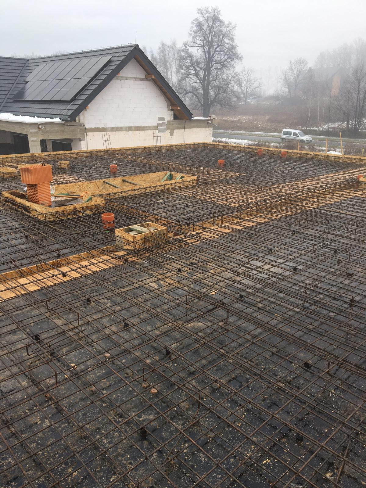 Siatka zbrojeniowa przygotowana na drewnianym szalunku, z widocznymi elementami instalacji i fragmentem budowanego domu z panelami słonecznymi w tle, sfotografowana w pochmurny dzień.