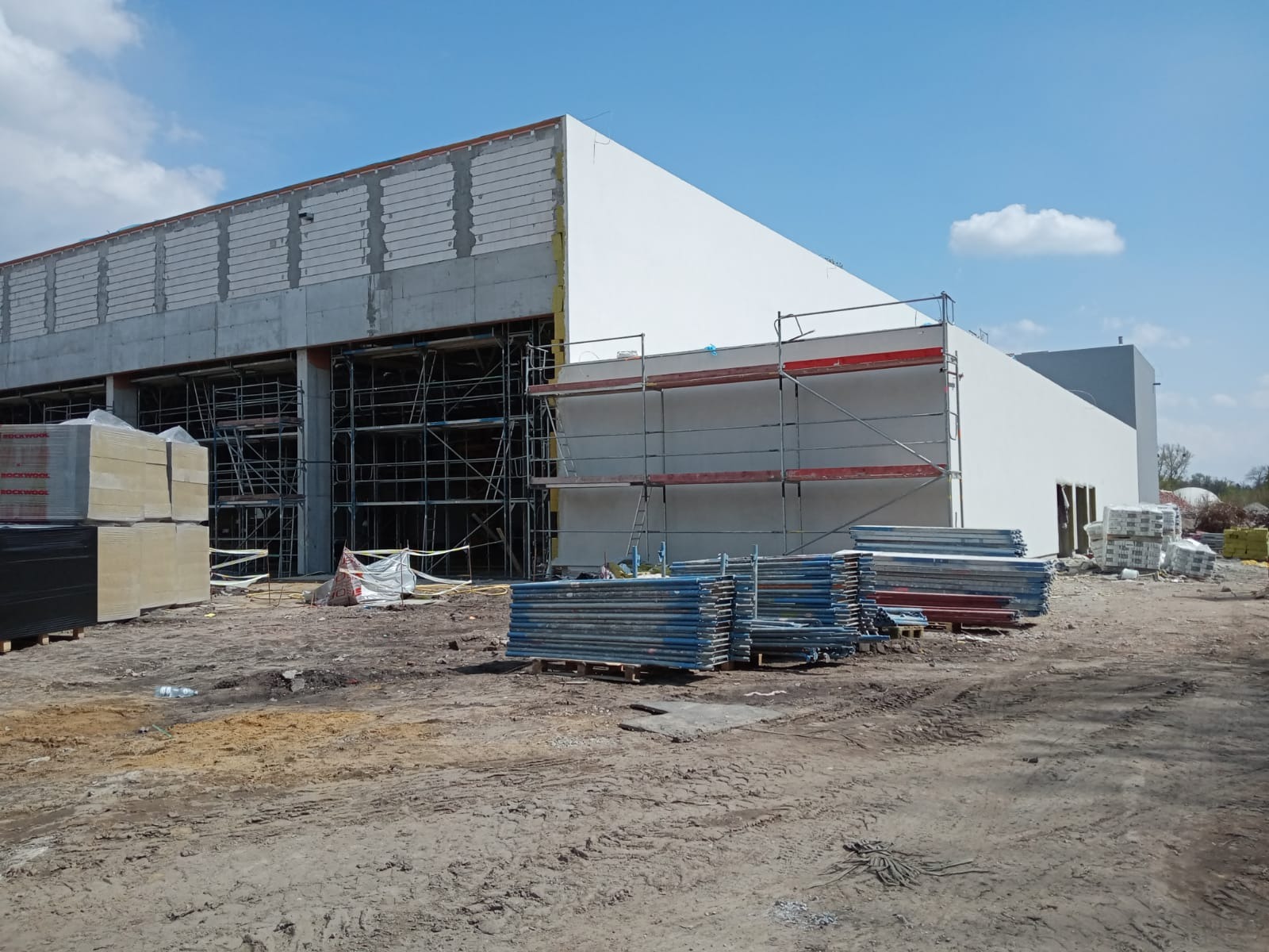 Budowa budynku murowanego z betonu komórkowego, z fragmentami ocieplenia i rusztowaniami, widoczne palety z materiałami budowlanymi i teren budowy.