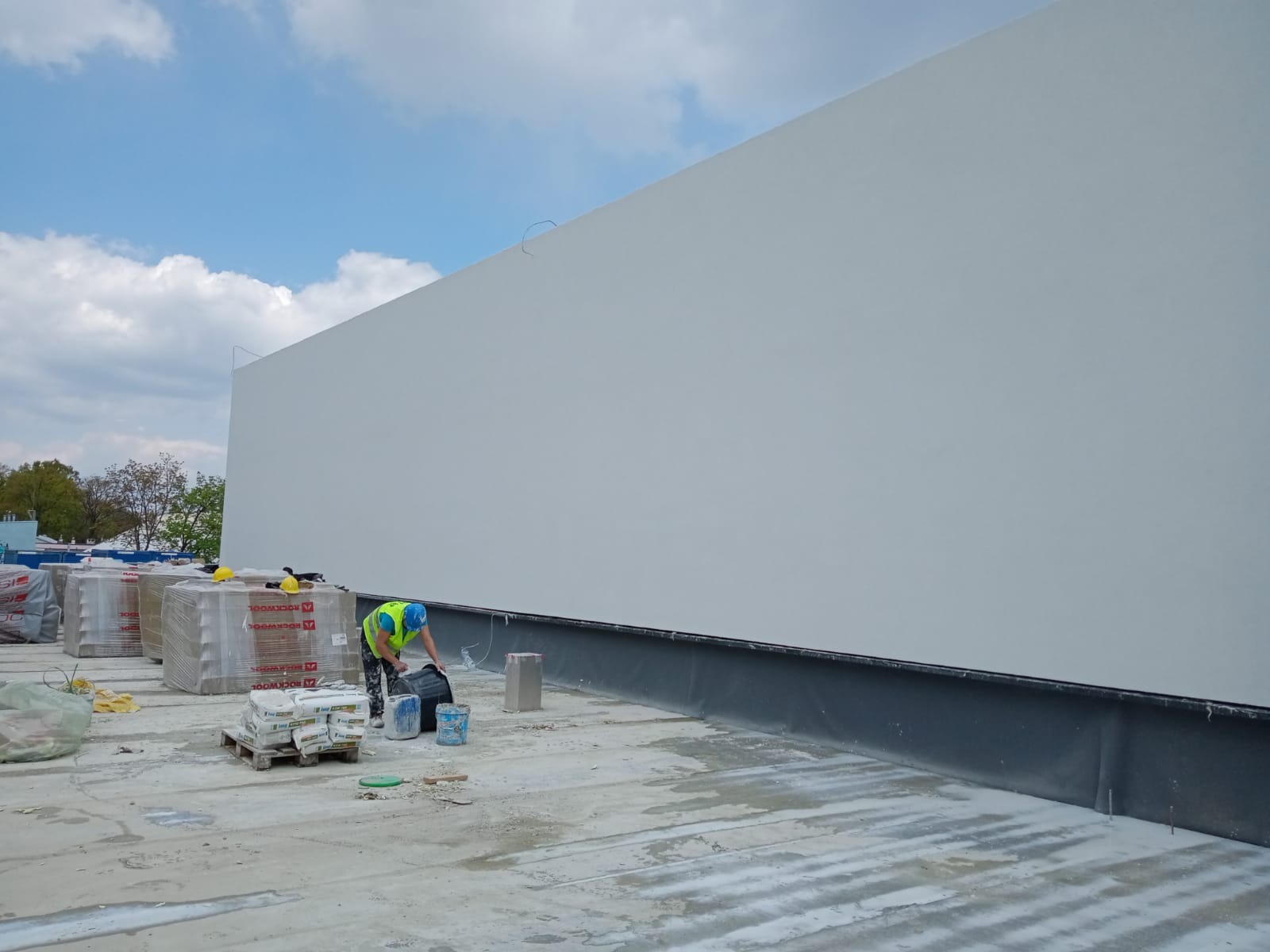 Pracownik w kamizelce odblaskowej miesza zaprawę na dachu budynku z ocieploną ścianą szczytową, widoczne paczki wełny mineralnej.