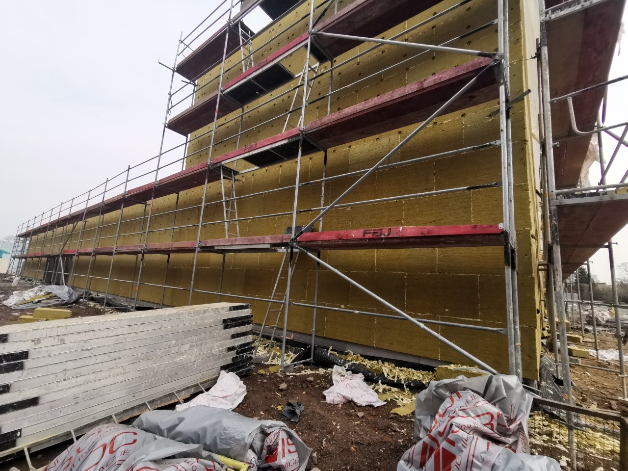 Budynek w trakcie ocieplania, widoczna warstwa żółtej izolacji na elewacji, rusztowanie i materiały budowlane składowane na ziemi.