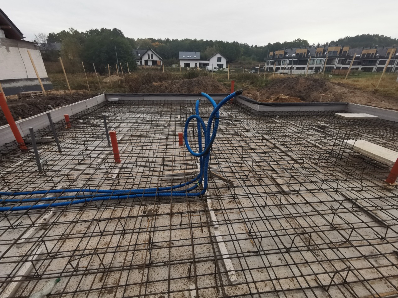 Zbrojenie płyty fundamentowej z wystającymi rurami instalacyjnymi w kolorze pomarańczowym i niebieskim, widok na budynki w tle.