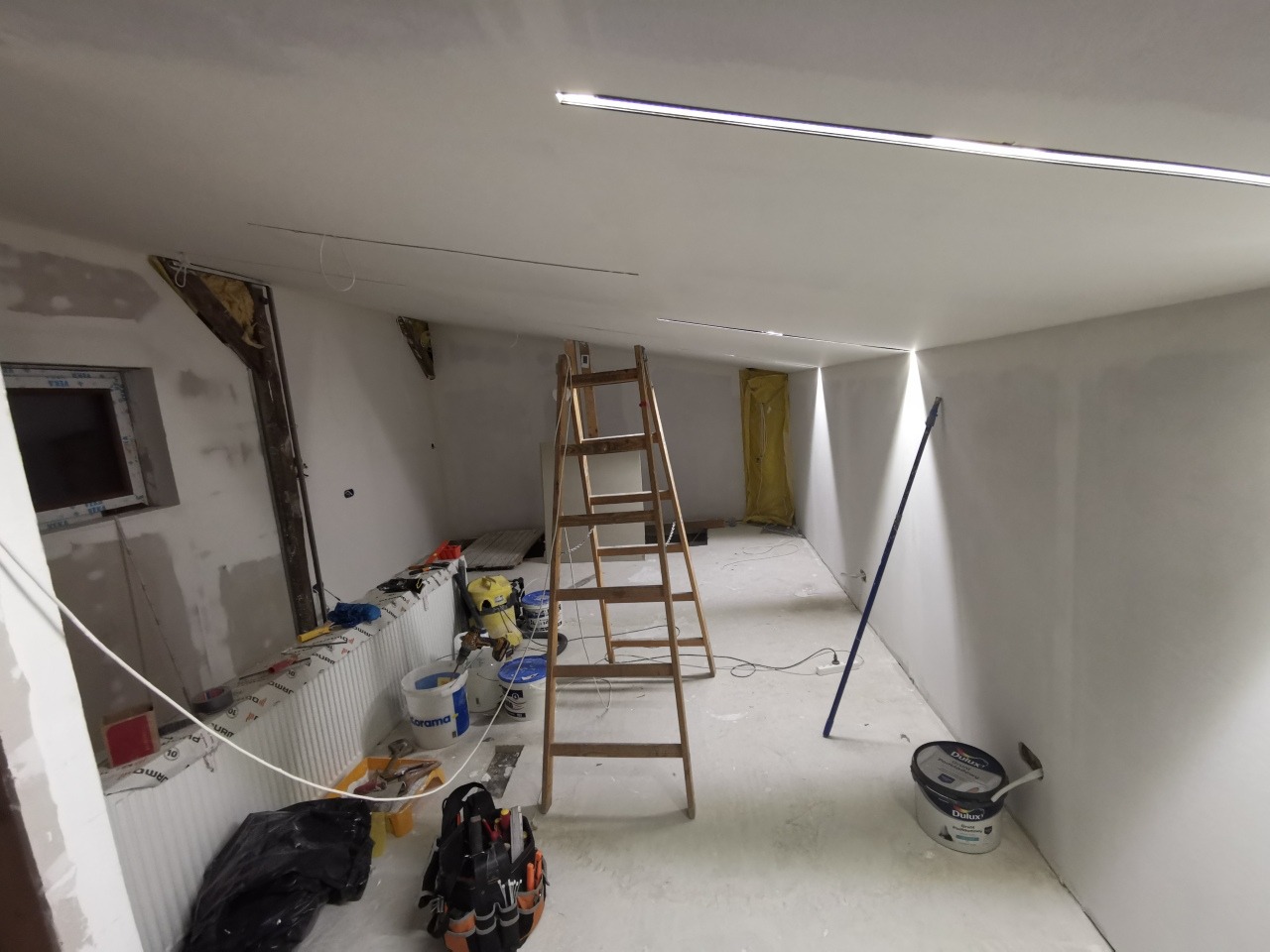Wnętrze w trakcie prac wykończeniowych: drewniana drabina, pędzle, wiaderka z farbą, narzędzia, widoczne listwy oświetleniowe na suficie, ściany przygotowane do malowania.