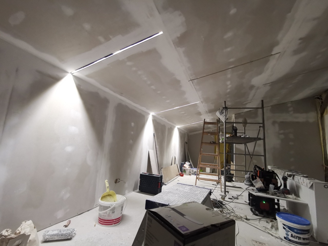 Wnętrze w trakcie wykańczania: ściany pokryte płytami gipsowo-kartonowymi, widoczne szpachlowanie i wnęki na oświetlenie liniowe, rusztowania w tle, narzędzia i materiały budowlane na podłodze.