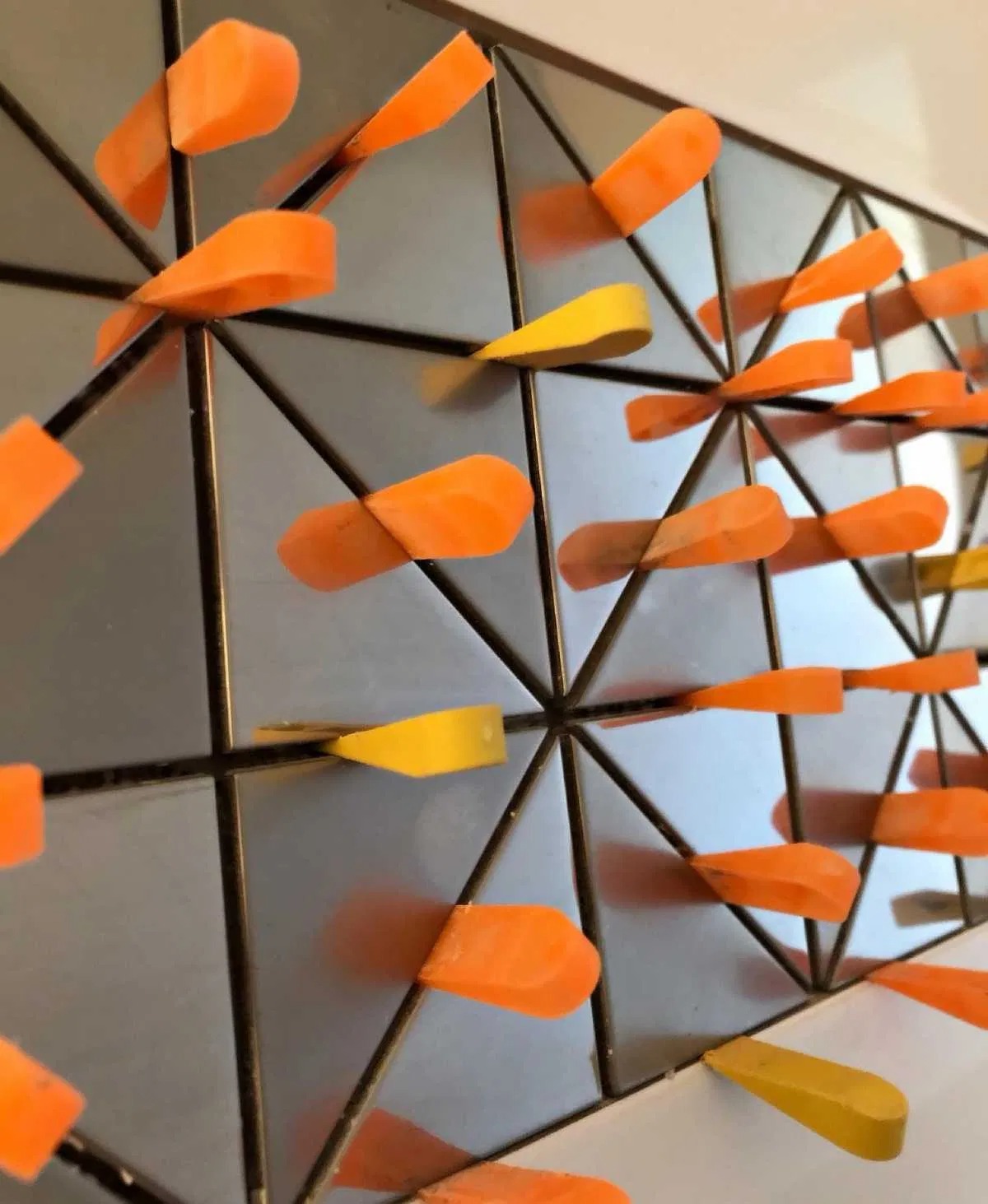 Abstrakcyjna dekoracja ścienna z geometrycznym wzorem, metalowa rama z trójkątami, pomarańczowe i żółte elementy dekoracyjne w kształcie strzałek, nowoczesny design.