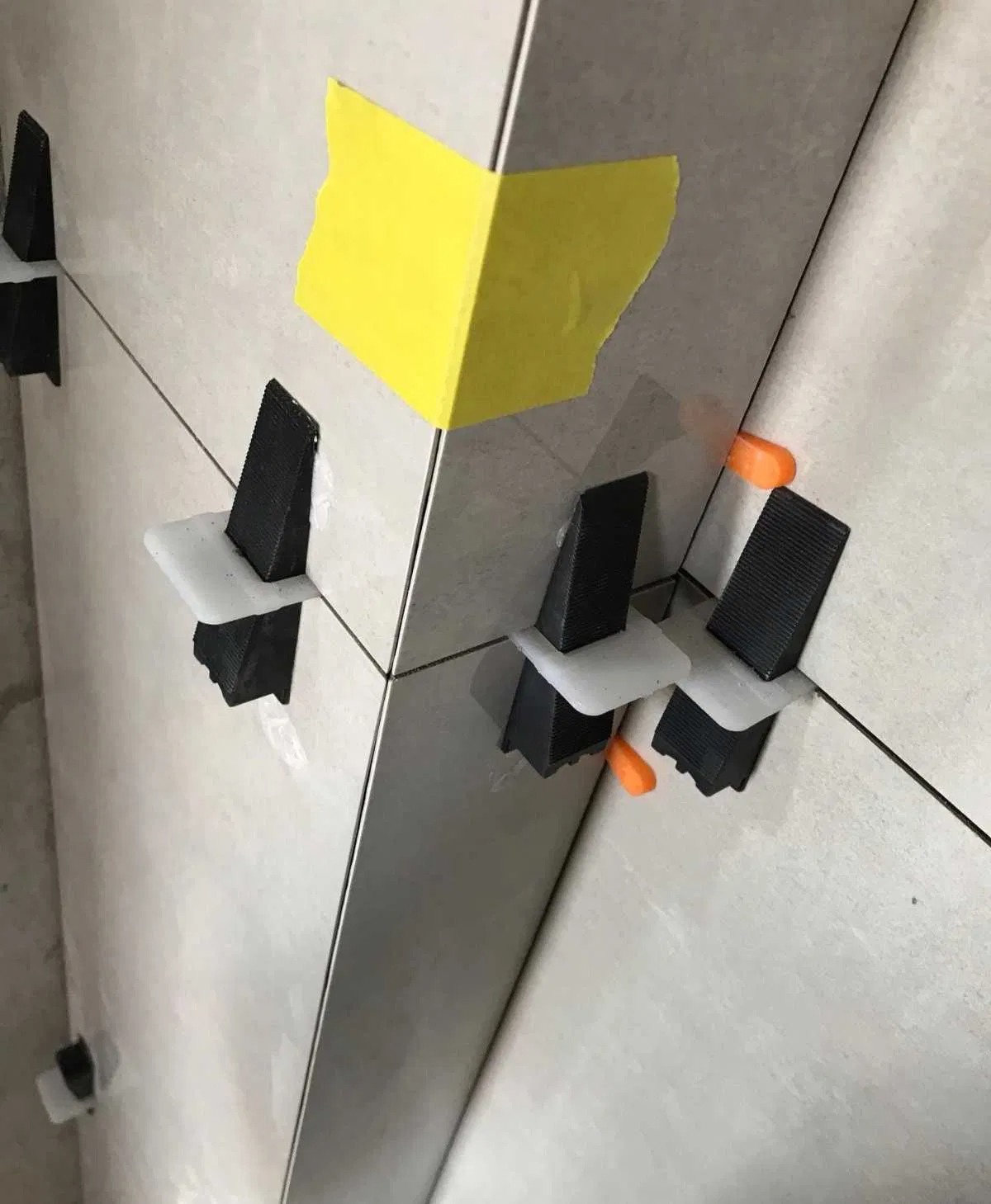 Układanie jasnych płytek ściennych z użyciem systemu poziomowania: czarne kliny i pomarańczowe dystanse, fragment ściany zabezpieczony żółtą taśmą malarską.