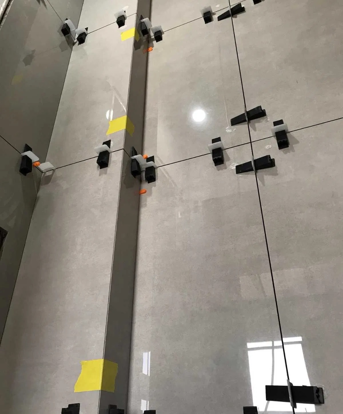 Układanie szarych płytek ściennych z użyciem systemu poziomowania płytek z czarnymi klipsami i żółtą taśmą maskującą, widoczne cienkie czarne krzyżyki dystansowe.