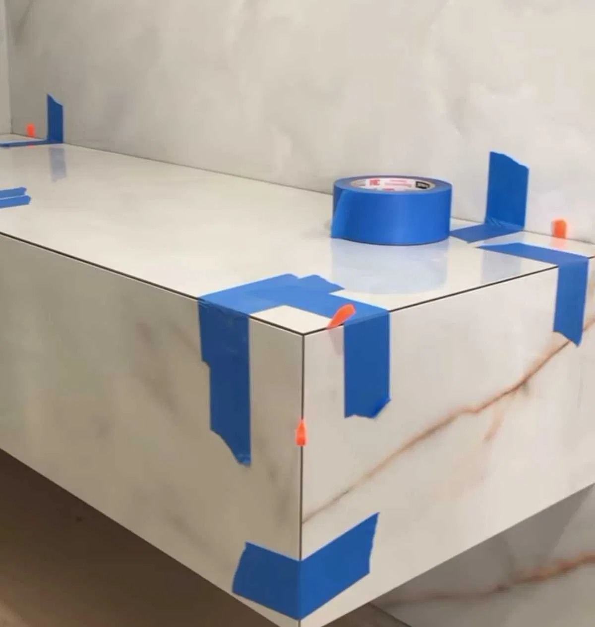 Biała, marmurkowa półka łazienkowa w trakcie wykańczania, zabezpieczona niebieską taśmą malarską i pomarańczowymi krzyżykami dystansowymi, z rolką taśmy na blacie.