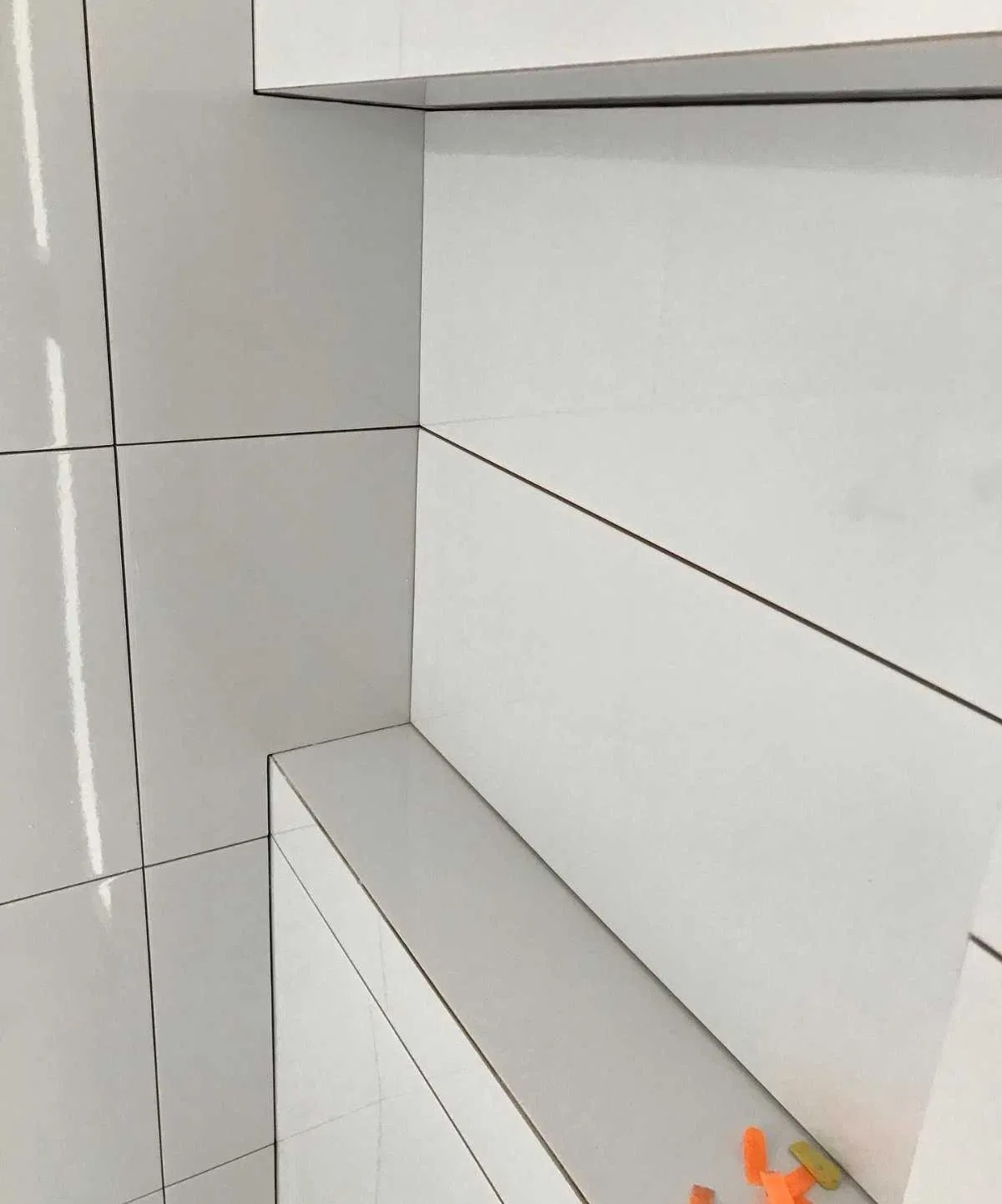 Świeżo ułożone, błyszczące, białe płytki ceramiczne z wąską fugą w rogu łazienki, widoczne poziomice i krzyżyki dystansowe.