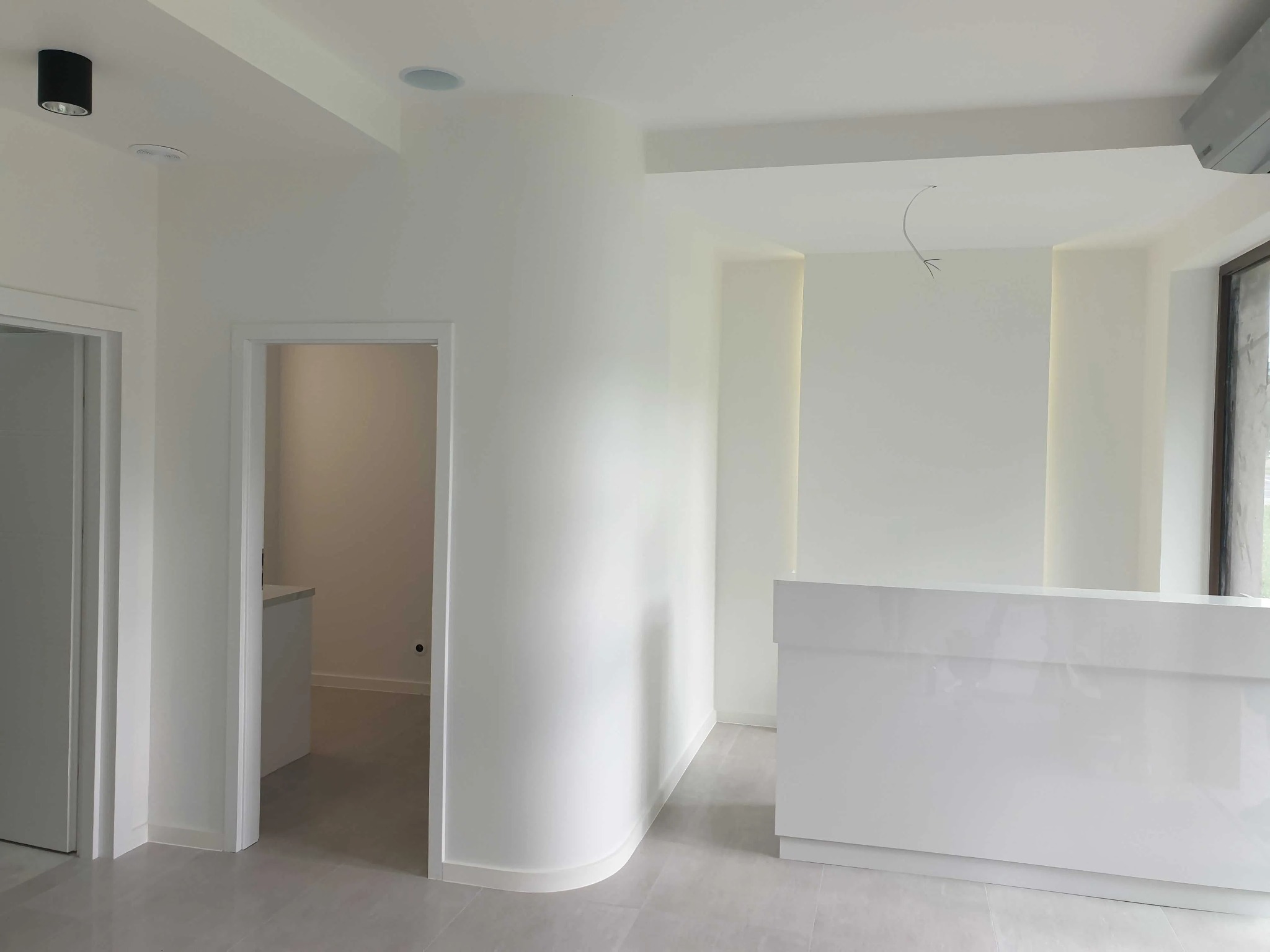 Nowoczesne, minimalistyczne wnętrze w Białystoku z białą, zaokrągloną ścianą, szarą podłogą i recepcją w połysku, widok na sąsiednie pomieszczenie.