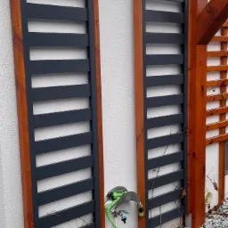 Dwa drewniane panele ogrodowe z poziomymi szczebelkami, przymocowane do białej ściany budynku, z widocznym systemem nawadniania z zielonym wężem i kamieniami dekoracyjnymi u podstawy.