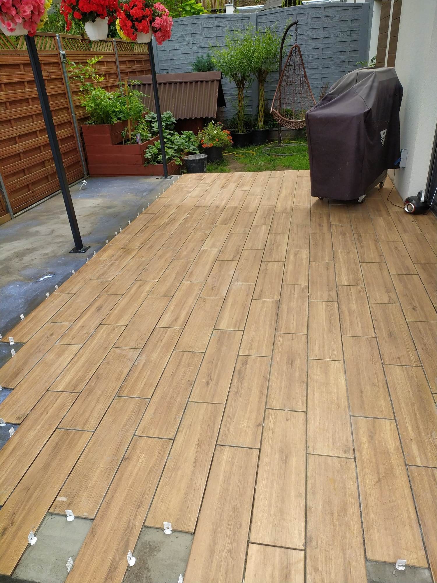 Układanie drewnopodobnych płytek na tarasie, widoczne plastikowe krzyżyki dystansowe, w tle roślinność ogrodowa i grill pod pokrowcem.