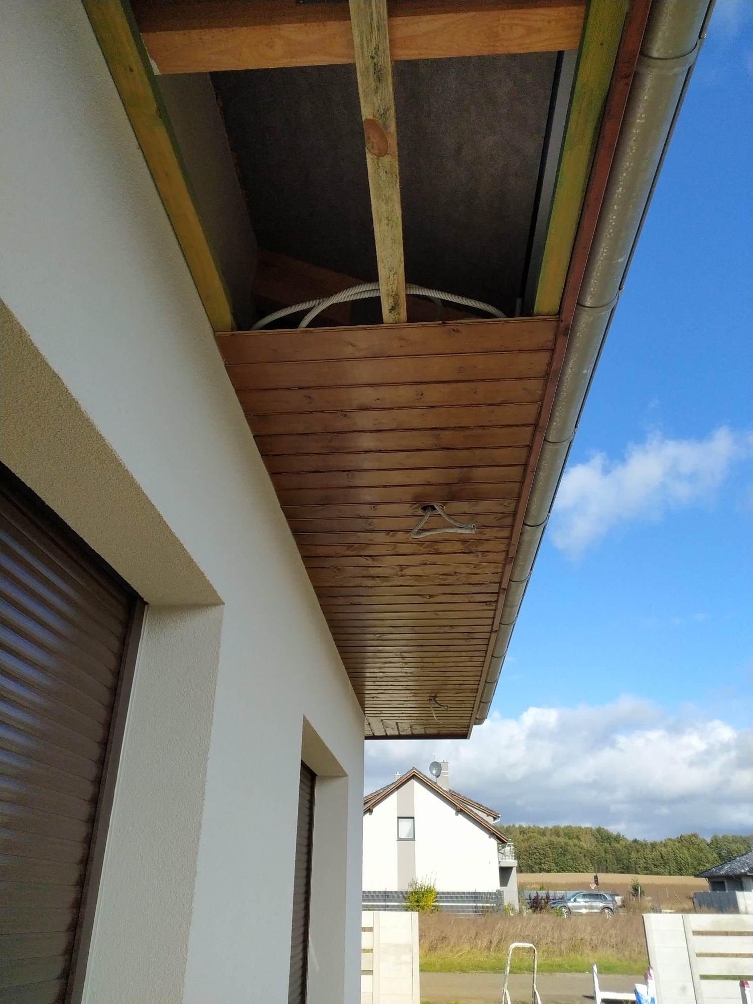 Detale wykończenia elewacji domu z drewnianą podbitką, widoczny fragment orynnowania, okna z roletami zewnętrznymi i tynkowana ściana.