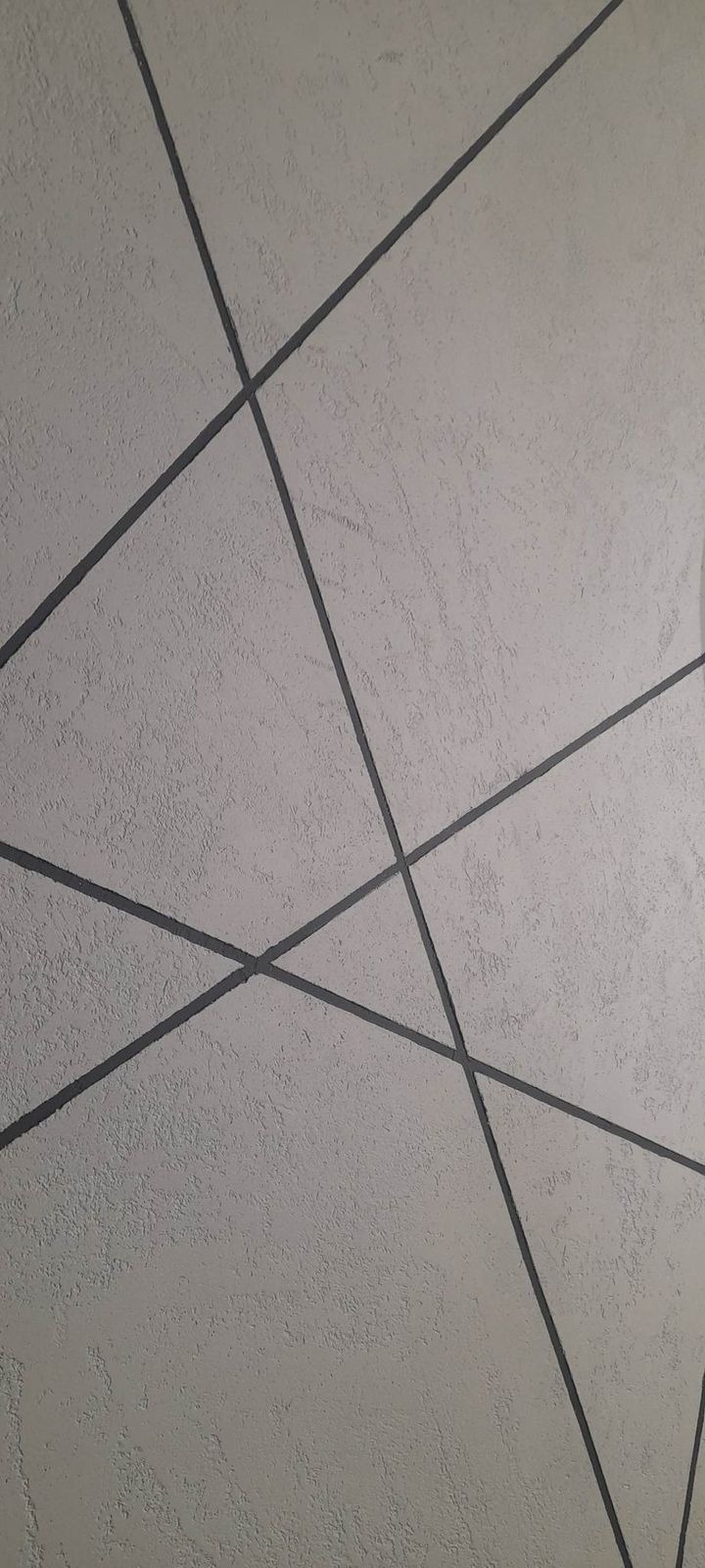 Dekoracyjna ściana z szarym tynkiem strukturalnym i geometrycznym wzorem z ciemnoszarych, ukośnych linii.