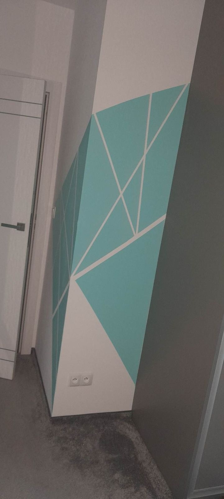 Ściana w Zielonej Górze z geometrycznym wzorem w odcieniach błękitu i bieli, wykonanym na białym tle, obok drzwi z srebrną klamką.