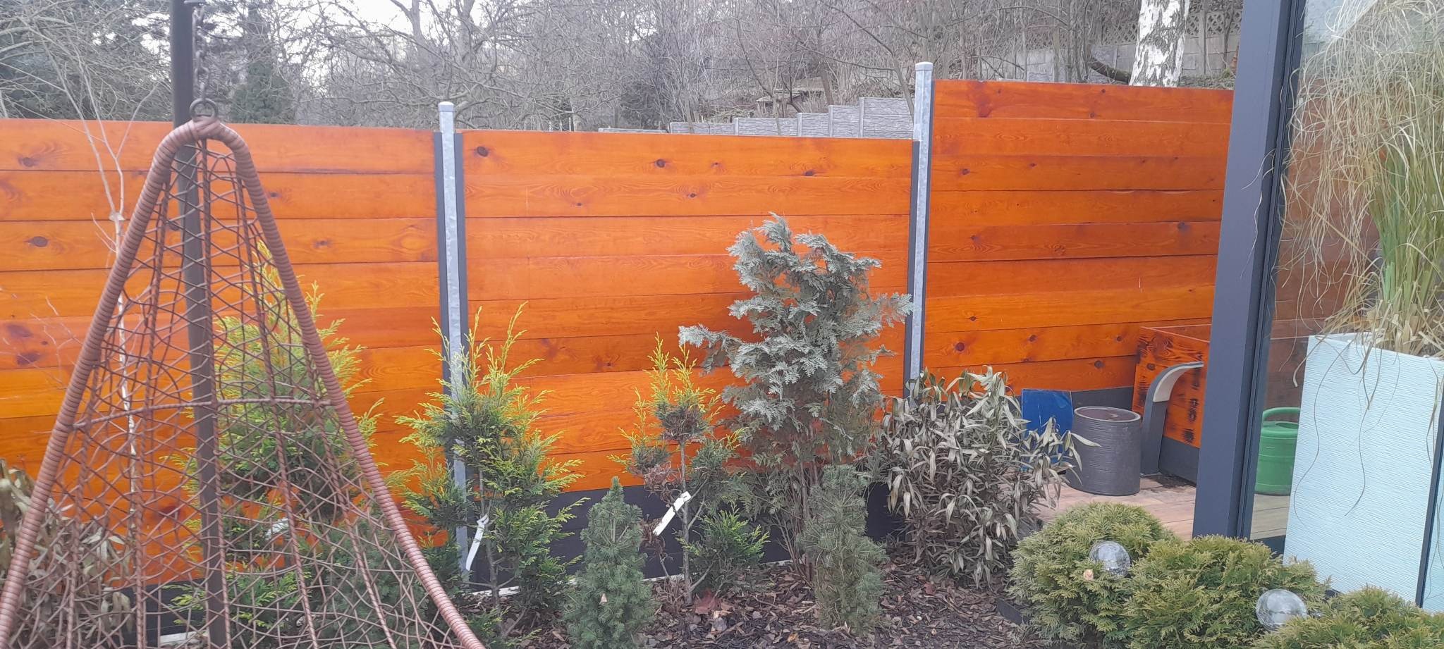 Nowoczesne ogrodzenie z drewnianych paneli o ciepłym, brązowym odcieniu, wsparte na metalowych słupkach, otoczone różnorodnymi krzewami iglastymi i liściastymi, z wiszącym fotelem kokonem po lewej...