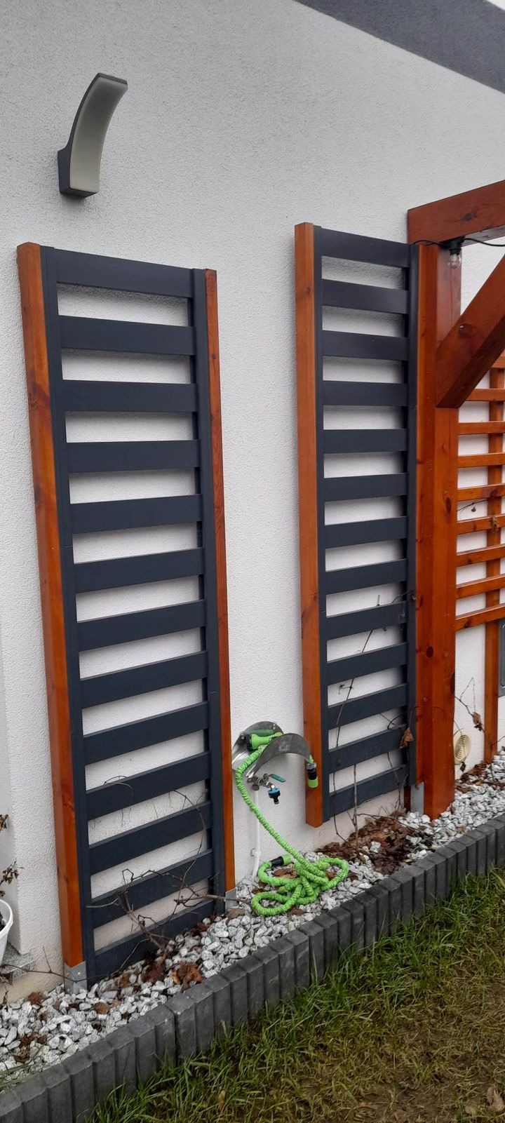 Dwa drewniane panele ogrodowe z poziomymi szczebelkami, przymocowane do białej ściany budynku, z widocznym systemem nawadniania z zielonym wężem i kamieniami dekoracyjnymi u podstawy.