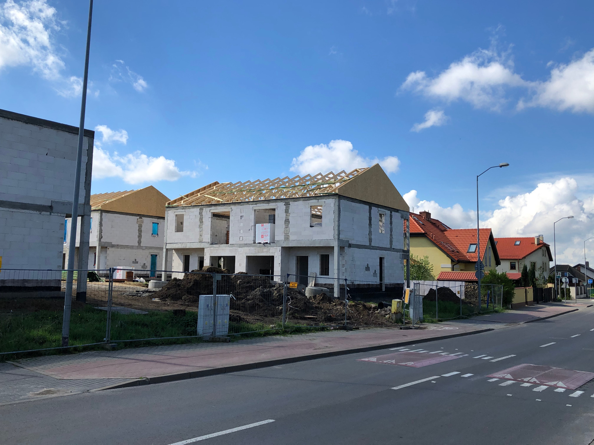 Wznoszone domy w stanie surowym otwartym z widoczną konstrukcją dachu i ścianami z białych bloczków, ogrodzone siatką budowlaną, na tle błękitnego nieba z chmurami.