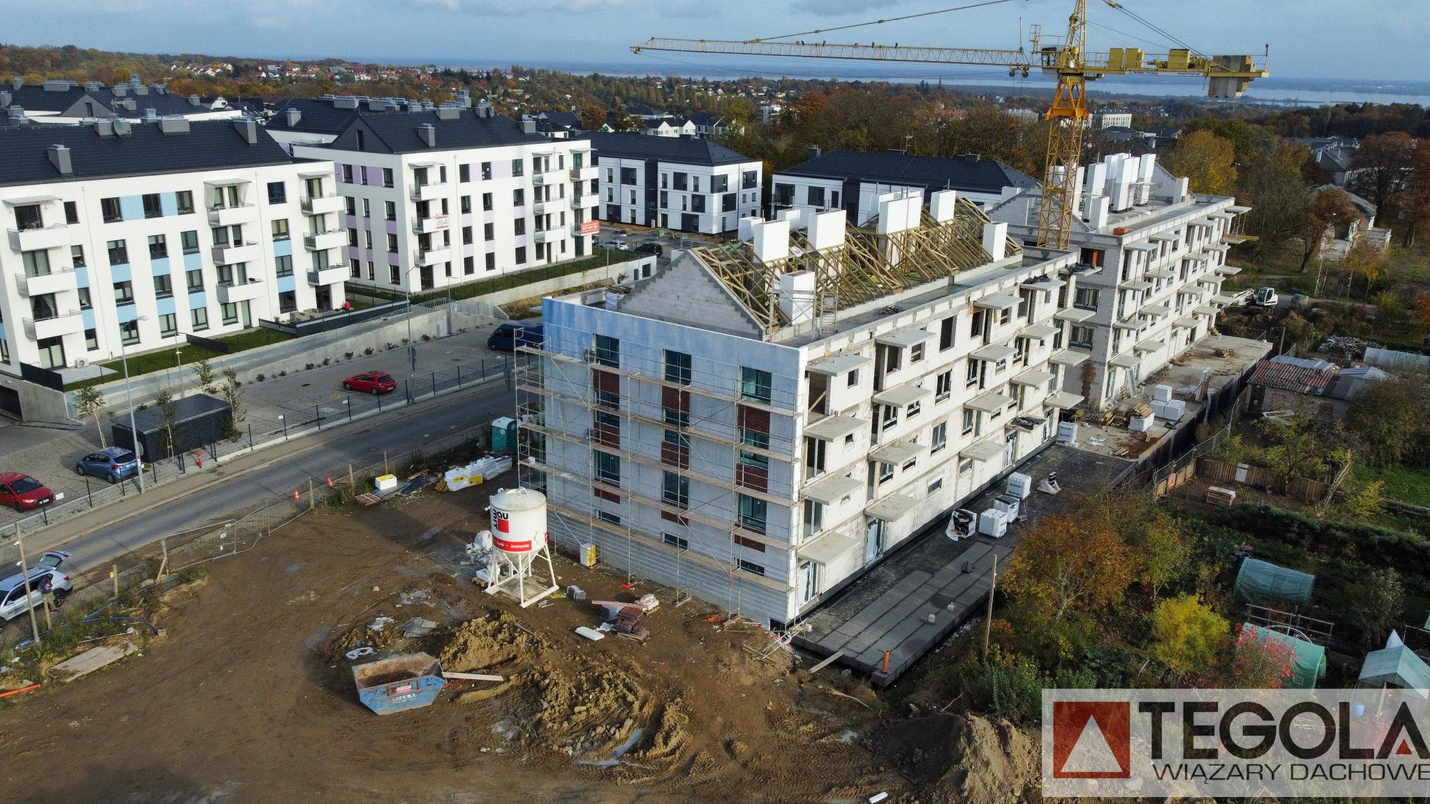 Wysoki kąt widoku na budowę wielopiętrowego budynku z białego betonu komórkowego, z widocznymi rusztowaniami, konstrukcją dachu w trakcie montażu, żółtym dźwigiem budowlanym w tle i okolicznymi...