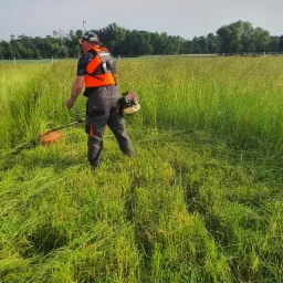 Pracownik w pomarańczowej kamizelce i szarych spodniach roboczych kosi wysoką trawę przy użyciu podkaszarki spalinowej na tle łąki i drzew.