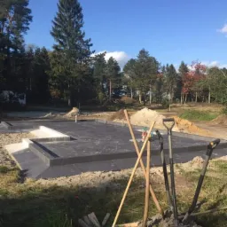 Przygotowany fundament pod budynek jednorodzinny, widoczne zbrojenie i zalane betonem ławy fundamentowe, na pierwszym planie narzędzia budowlane: łopaty i grabie wbite w ziemię.