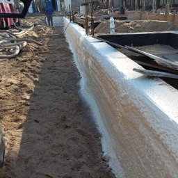 K&D-Izolacje KAMIL KOWALSKI - Świeżo natryśnięta, jasna pianka izolacyjna na zewnętrznej ścianie fundamentu w trakcie budowy, widoczny fragment opony pojazdu i przewody robocze.