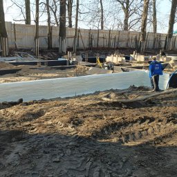 K&D-Izolacje KAMIL KOWALSKI - Świeżo zaizolowane fundamenty budynku z widoczną strukturą piany izolacyjnej, pracownik w niebieskiej kurtce z logo firmy na placu budowy, w tle ogrodzenie i drzewa.