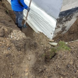 K&D-Izolacje KAMIL KOWALSKI - Pracownik w niebieskiej bluzie z logo firmy zasypuje ziemią fundament ocieplony pianką PUR i zabezpieczony folią kubełkową, widoczny drenaż.