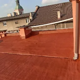 Czerwony, płaski dach pokryty nową izolacją bitumiczną, widok z góry na tle zabudowy miejskiej z kościelną wieżą.