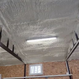 Poddasze ocieplone pianką PUR, widoczne krokwie i lampa oświetlająca nierównomierną strukturę pianki, ściany wykończone płytą OSB z małym oknem.