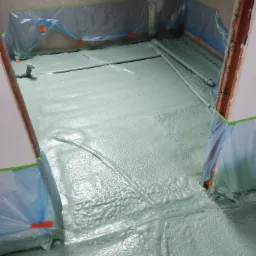 Świeżo wylana, jasnoszara wylewka na podłodze w pomieszczeniu, zabezpieczona folią i taśmą malarską przy ścianach, z widocznymi rurami grzewczymi w podłodze i zaworem przy ścianie.