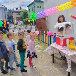 Dzieci ustawione w kolejce przed stoiskiem z popcornem, obsługiwanym przez uśmiechniętą kobietę; kolorowe dekoracje i ogrodzenie w tle.