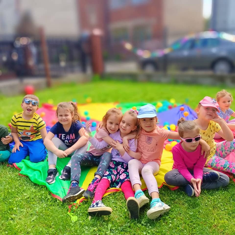 Grupa uśmiechniętych dzieci siedzi na kolorowej macie na trawie, w tle rozrzucone kolorowe piłki i zamazane budynki.