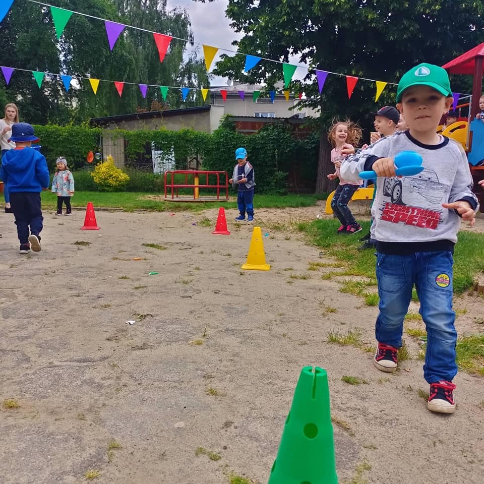 Chłopiec w zielonej czapce i bluzie z samochodem, bawiący się niebieską łopatką na placu zabaw z kolorowymi pachołkami i girlandami.