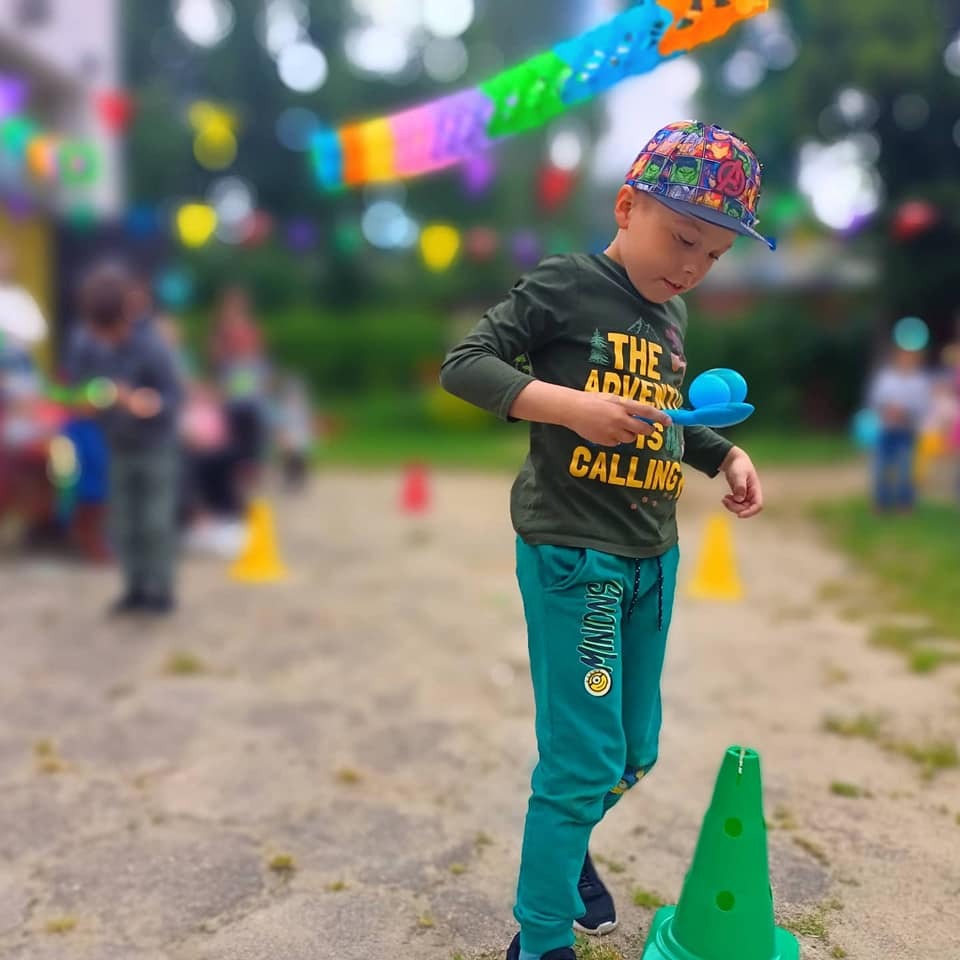 Chłopiec w czapce z motywem superbohaterów przenosi niebieską piłeczkę na łyżce, balansując nad zielonym pachołkiem podczas zabawy na zewnątrz.