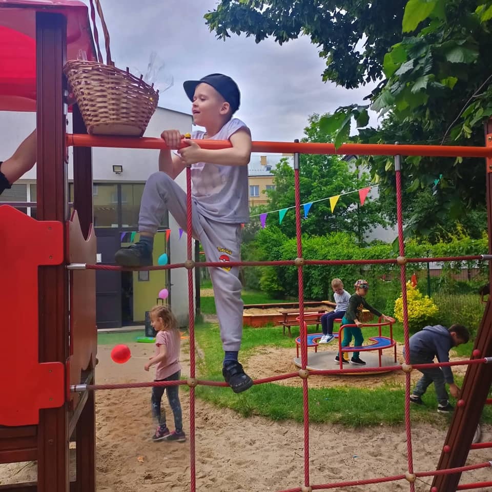 Chłopiec w czapce wspina się po czerwonej konstrukcji linowej na placu zabaw, obok kosz z zabawkami, w tle dzieci bawiące się na karuzeli i w piaskownicy, dekoracje z kolorowych chorągiewek.