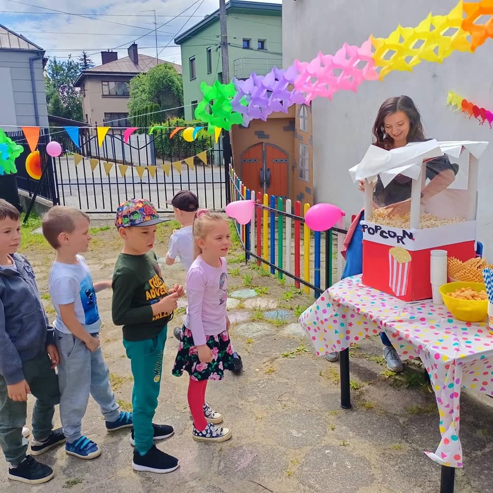 Dzieci ustawione w kolejce przed stoiskiem z popcornem, obsługiwanym przez uśmiechniętą kobietę; kolorowe dekoracje i ogrodzenie w tle.