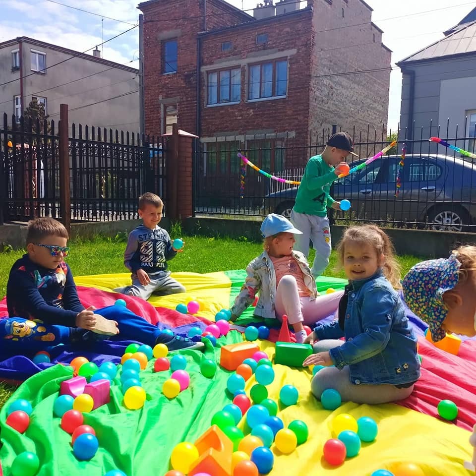 Grupa dzieci bawi się na kolorowej chuście animacyjnej z piłeczkami i miękkimi klockami na trawie przed budynkiem w żłobku.