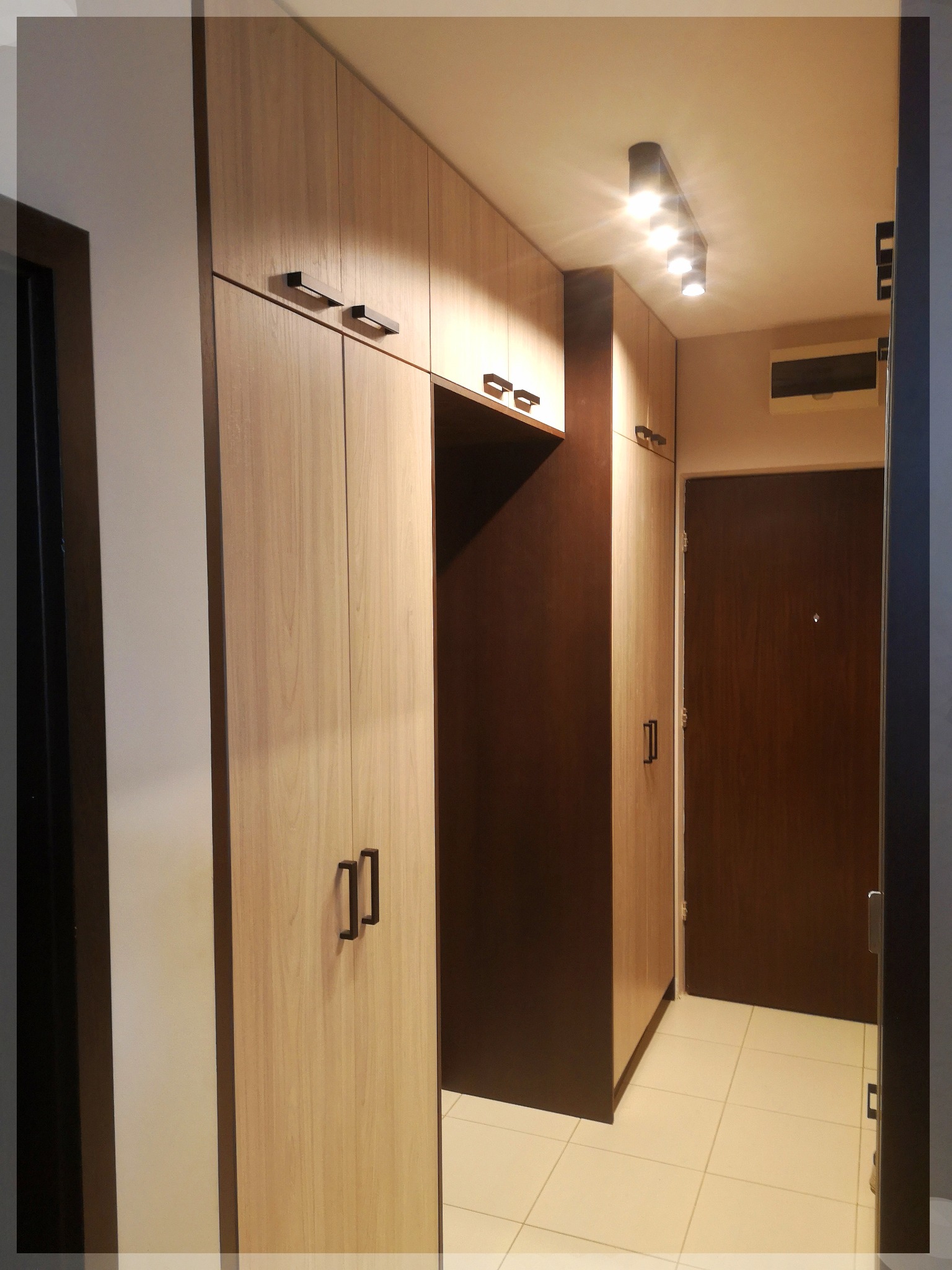 Zabudowa przedpokoju z szafami o jasnym, drewnopodobnym wykończeniu i ciemnymi uchwytami, zintegrowana z ciemnobrązową wnęką i drzwiami wejściowymi.