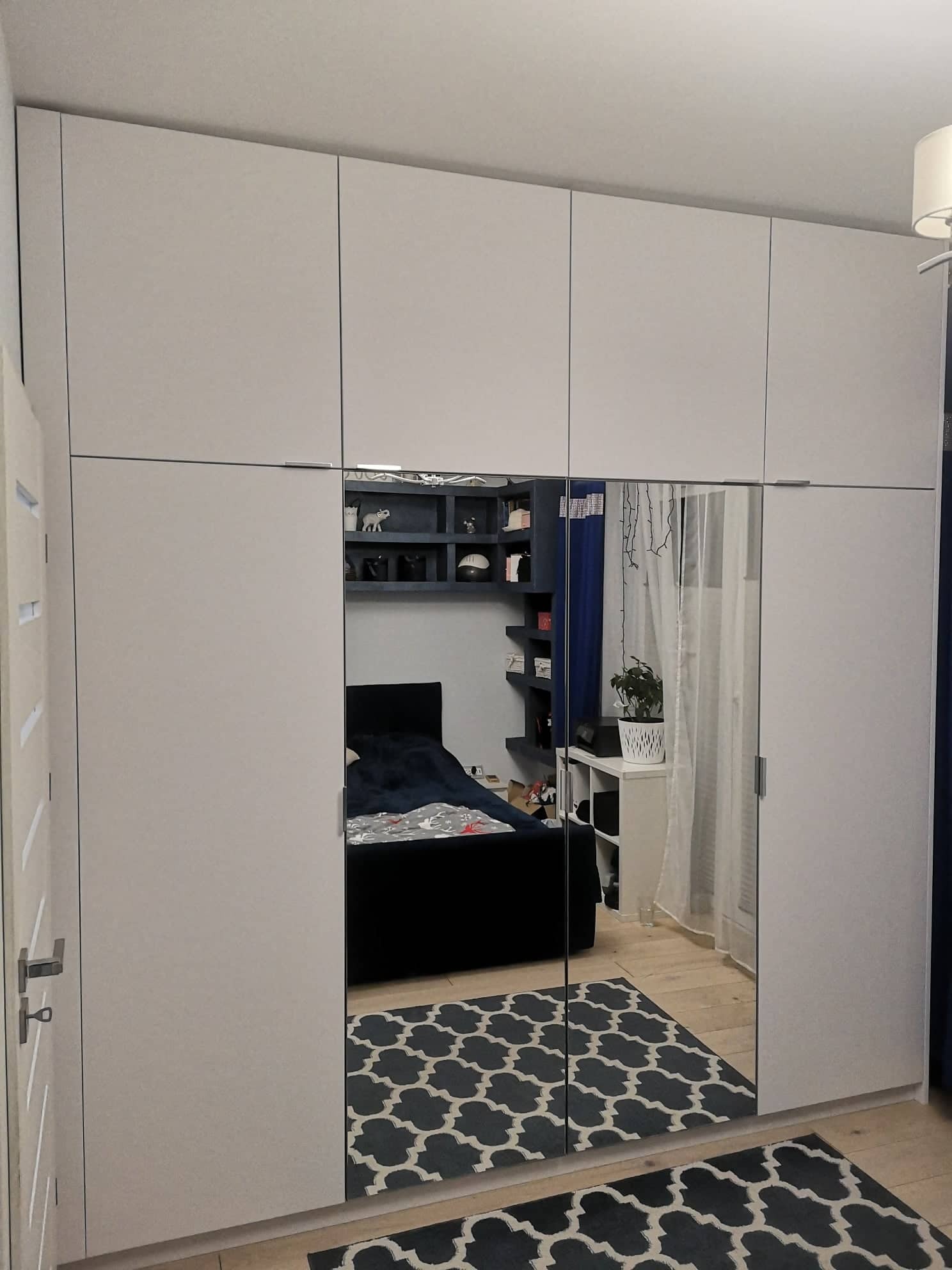 Szafa z lustrzanymi drzwiami przesuwnymi, odbijająca wnętrze sypialni z łóżkiem i regałami na ścianie, na podłodze wzorzysty dywan.