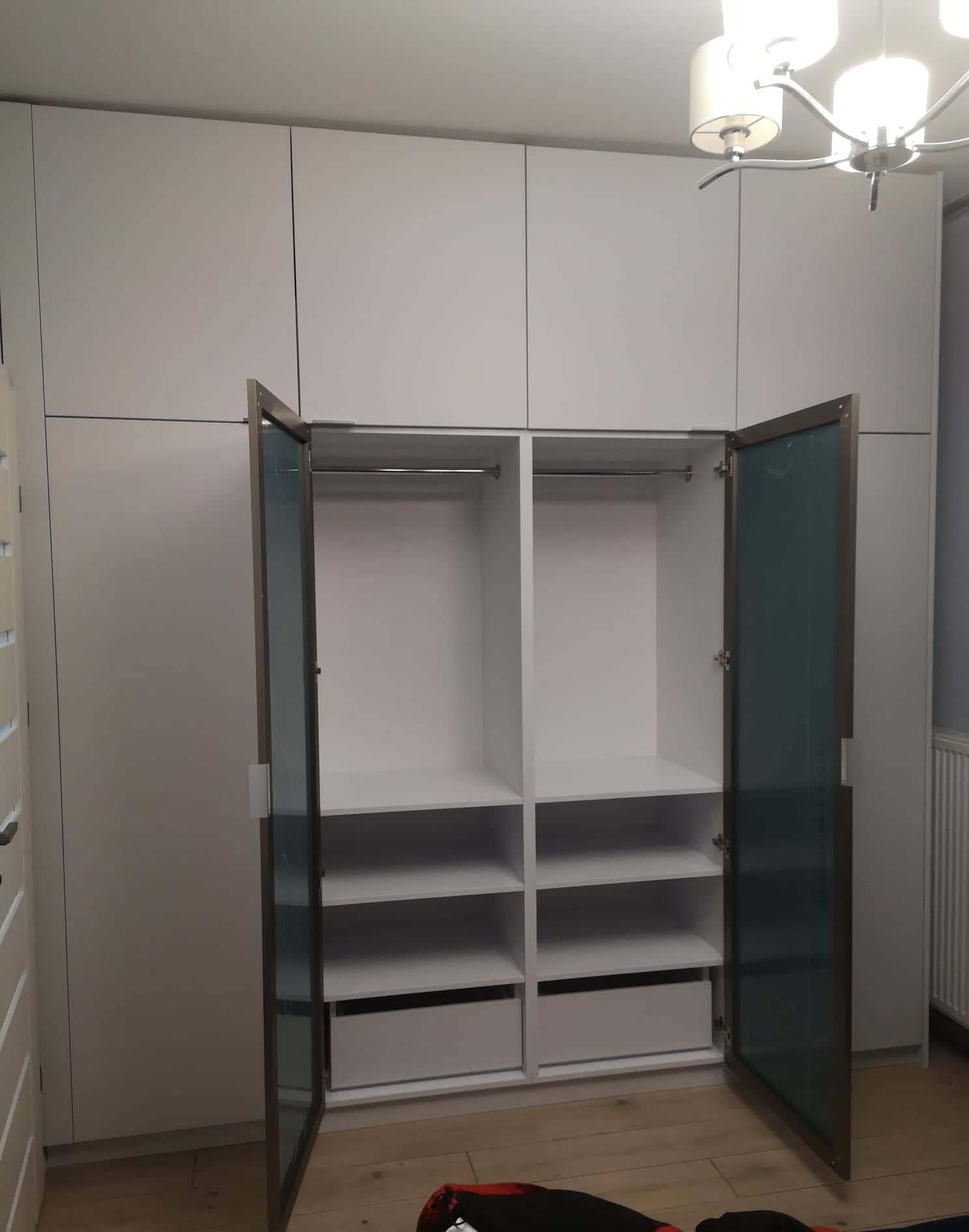 Biała szafa wnękowa z przesuwnymi drzwiami z szarym obramowaniem i półkami, częściowo otwarta, widoczne szuflady, minimalistyczny design, oświetlenie sufitowe w tle.